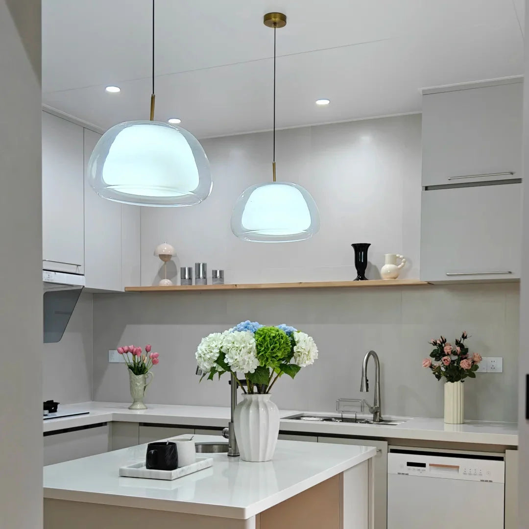 Curved glass pendant light with clear bell shade, warm light reflecting in kitchen setting.