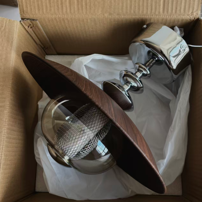 A ceiling fan light kit with a dark brown wood-grain blade, chrome metal mounting hardware, and a clear glass shade, shown inside its cardboard box with protective white paper.