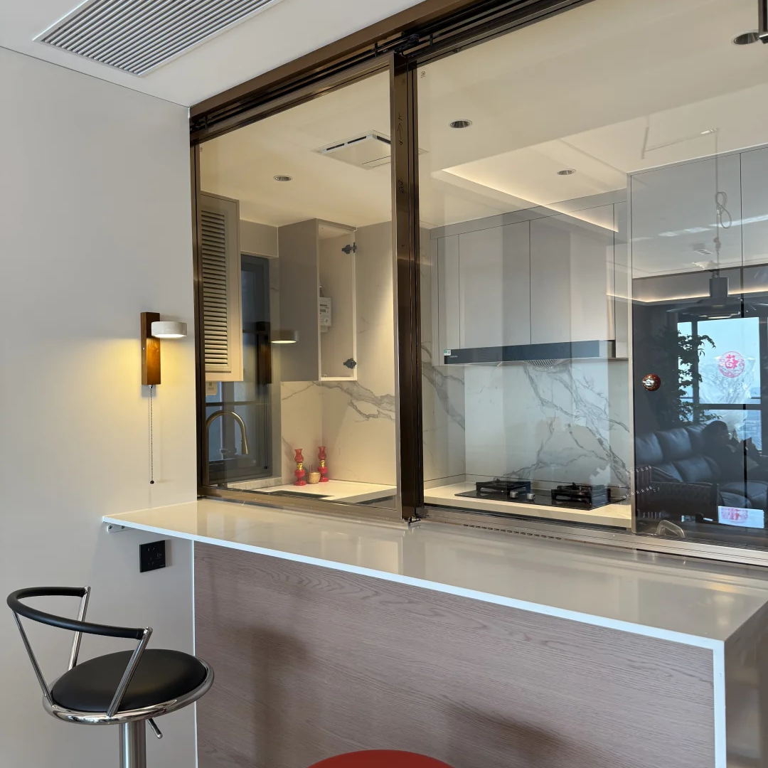 Modern kitchen interior with a glass partition and counter, featuring a white countertop, light wood base, a black bar stool, and a red stool in the foreground.
