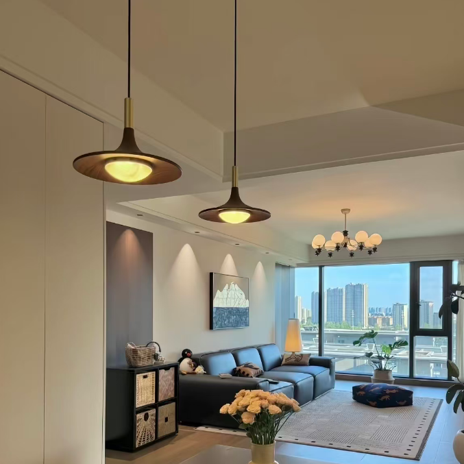 A modern living room interior with two pendant lights hanging over a dining table, a dark blue sofa in the background, large windows with a city view, and a vase of flowers on the table.