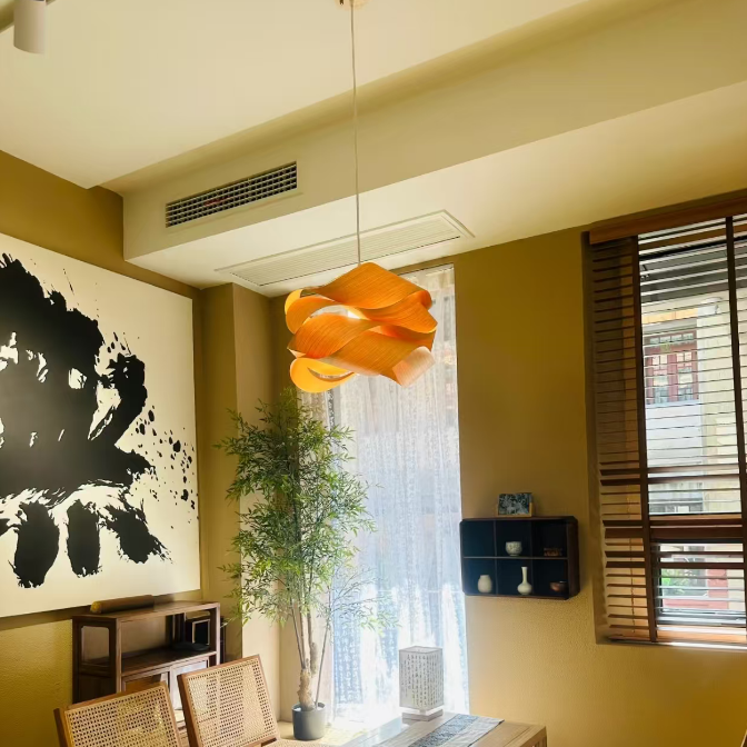 A modern Japanese-style living room featuring a distinctive wavy orange paper lantern pendant light hanging over a low wooden table. The room has warm beige walls, tatami-style flooring, woven cane chairs, and a large black ink brushstroke wall art piece.