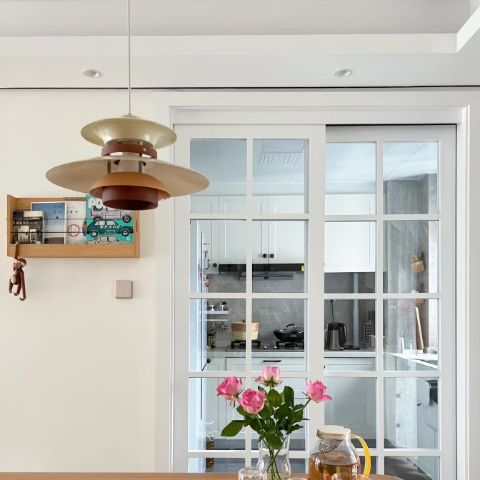 A modern pendant light with layered metallic gold and bronze shades hangs over a wooden dining table in a bright, contemporary dining room. A wooden tray on the table holds pink roses and glassware, with white-framed glass sliding doors in the background revealing a kitchen.