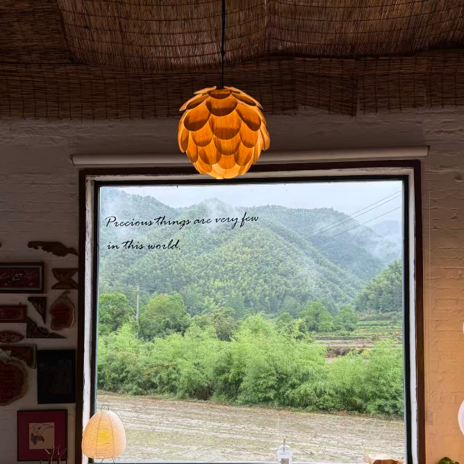 A cozy interior scene with a large window overlooking a lush green mountain landscape. A distinctive orange-yellow layered pendant light hangs from the ceiling, and the window has the text 'Previous things are very few in this world' written on it.