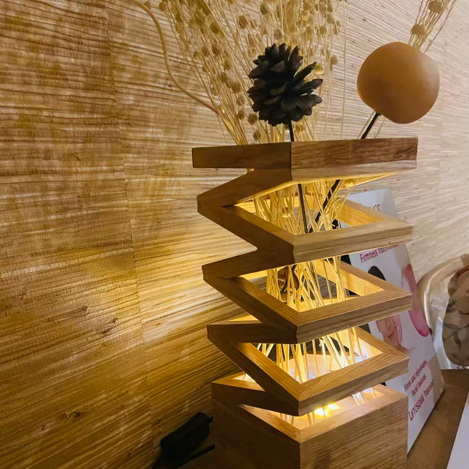 A modern wooden zigzag table lamp with warm white LED lights glowing inside, styled with dried grass, a pinecone, and a round decorative piece on top, set against a textured bamboo wall.