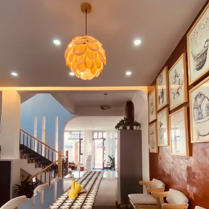 A modern interior space with a prominent orange flower-shaped pendant light hanging from a white ceiling. The room features a long black-and-white geometric patterned runner rug, a dark wooden dining table, several light wood chairs with cream cushions, a staircase on the left, and framed artwork on a terracotta-colored wall on the right.