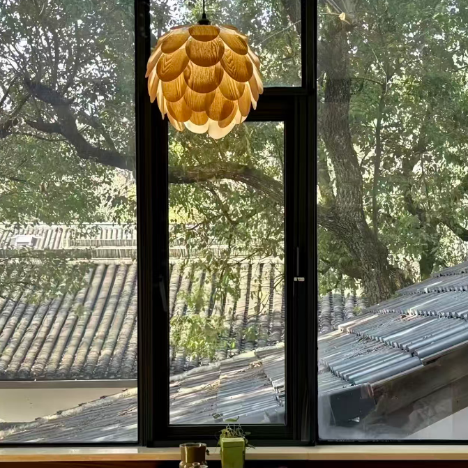 A modern pendant light with a layered, lotus-like golden shade hangs in front of a large black-framed window. Outside, trees and traditional tiled roofs are visible, while a small wooden table sits below the window in a styled interior setting.