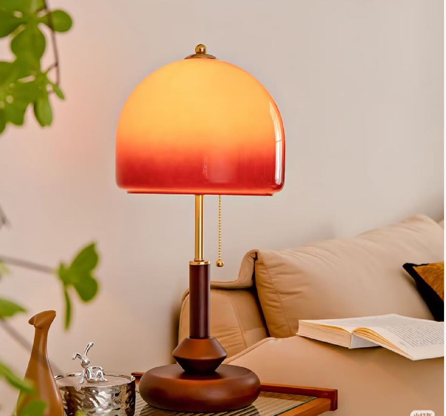 A table lamp with a warm gradient glass shade fading from yellow to orange to red, set on a side table next to a beige sofa with an open book nearby.