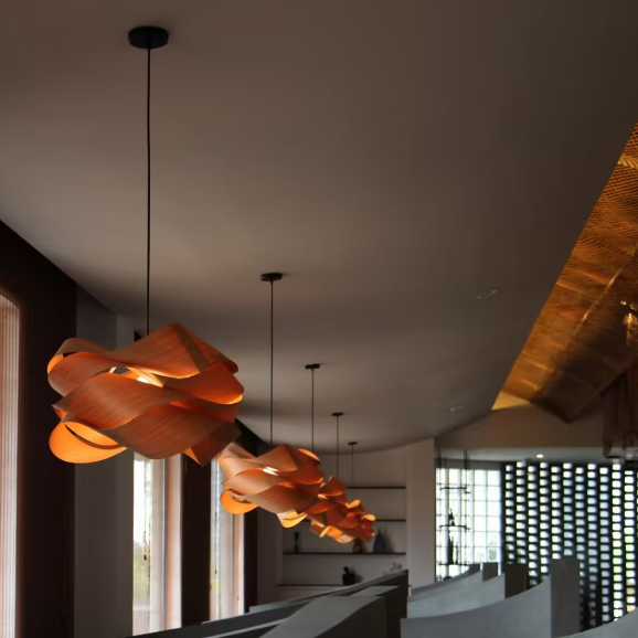 A row of modern pendant lights with layered, petal-like wooden shades in a warm orange-brown tone, hanging over curved concrete seating in a minimalist interior space.