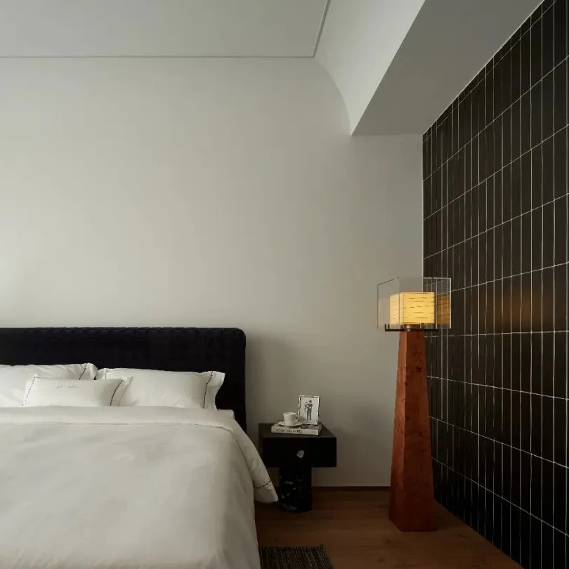 A modern bedroom interior featuring a neatly made bed with white bedding and a dark headboard, set against a white wall next to a black tiled accent wall with a lit floor lamp.