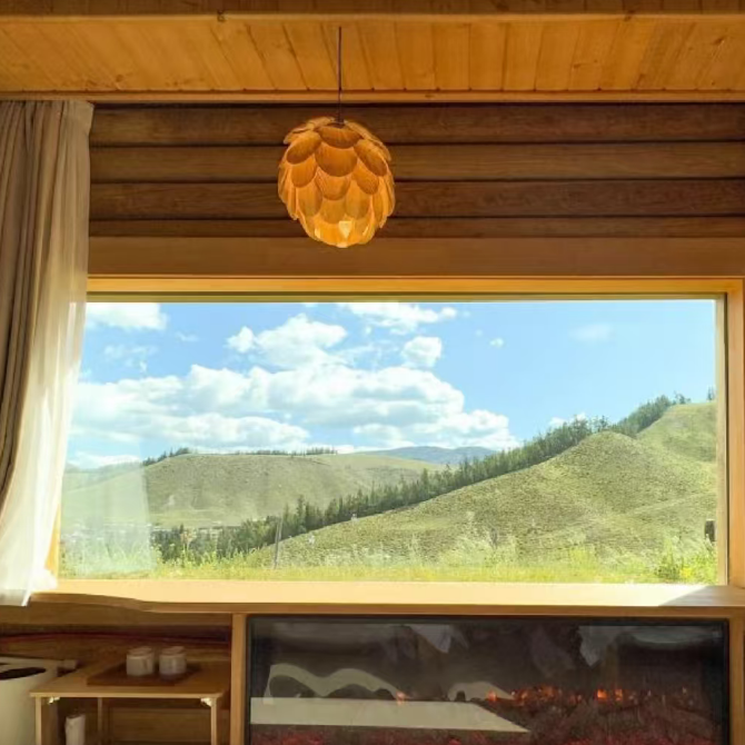 A modern interior room with a large floor-to-ceiling window overlooking green hills under a blue sky. A wood-burning fireplace is built into the window sill below, with a wooden ceiling, hanging pendant light, bench, and neutral textiles visible in the room.