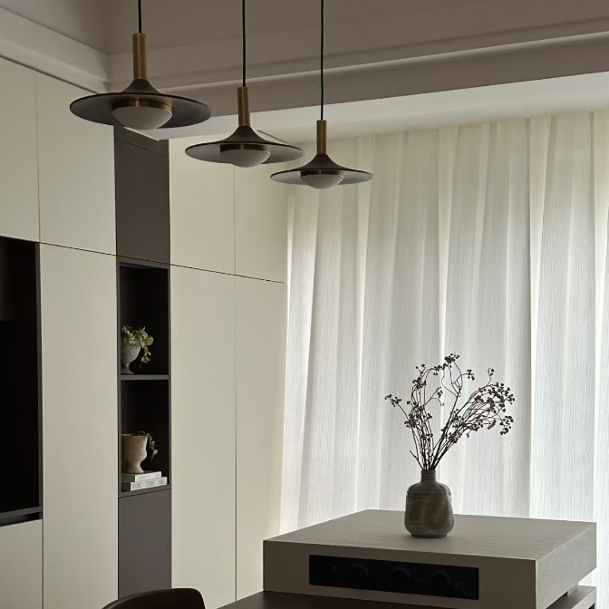 A modern interior scene with a dark wood dining table, a white pedestal table holding a vase of dried branches, built-in cream cabinets, pendant lights, and sheer white curtains.