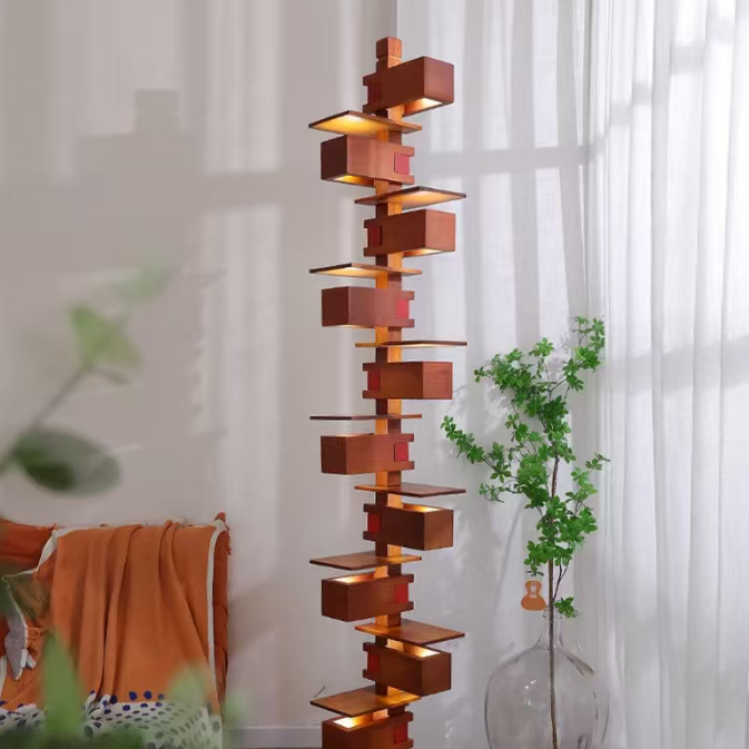 A tall, modern geometric floor lamp made of stacked reddish-brown wooden boxes and shelves with warm built-in lighting, standing in a bright room next to a window with white curtains and a glass vase with greenery.