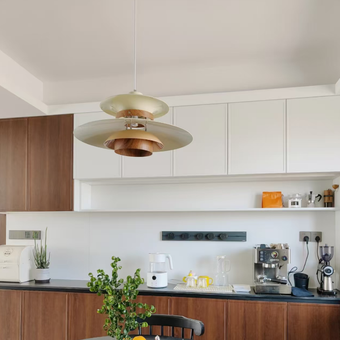 A modern kitchen interior with white cabinets, dark wood lower cabinets, a black countertop, and a hanging pendant light with layered metallic shades. A round black dining table with chairs is in the foreground, along with a potted plant, open book, tissue box, and small kitchen appliances.