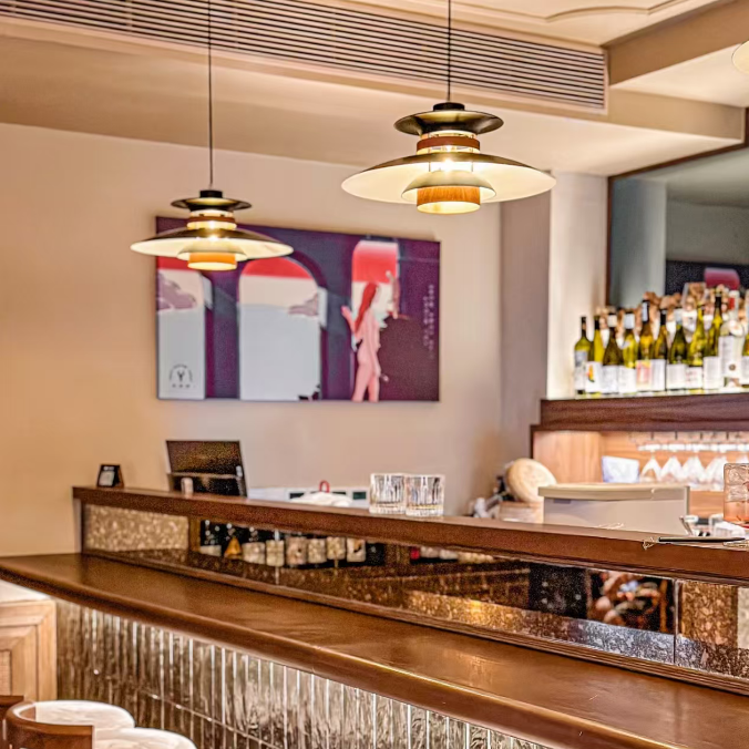 A modern bar interior with a wooden counter, textured glass tile front, white upholstered stools, pendant lights, and a framed artwork on the wall.