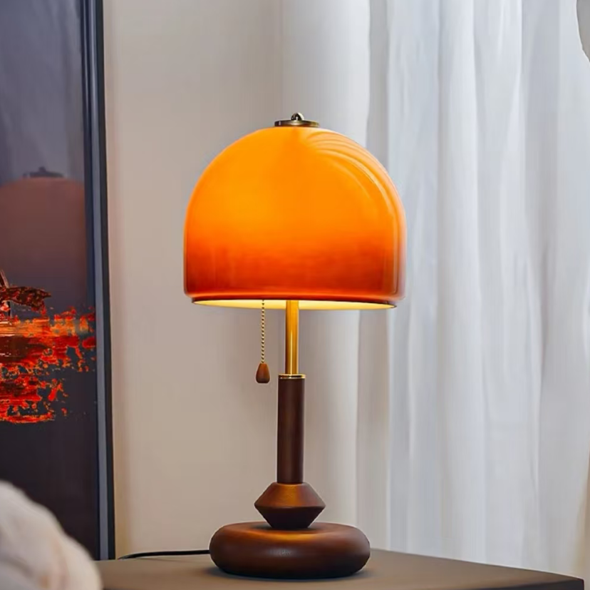 A table lamp with a glowing orange dome shade, a dark brown wooden stem, and a round dark base, placed on a tabletop in a room with white curtains and a framed artwork in the background.