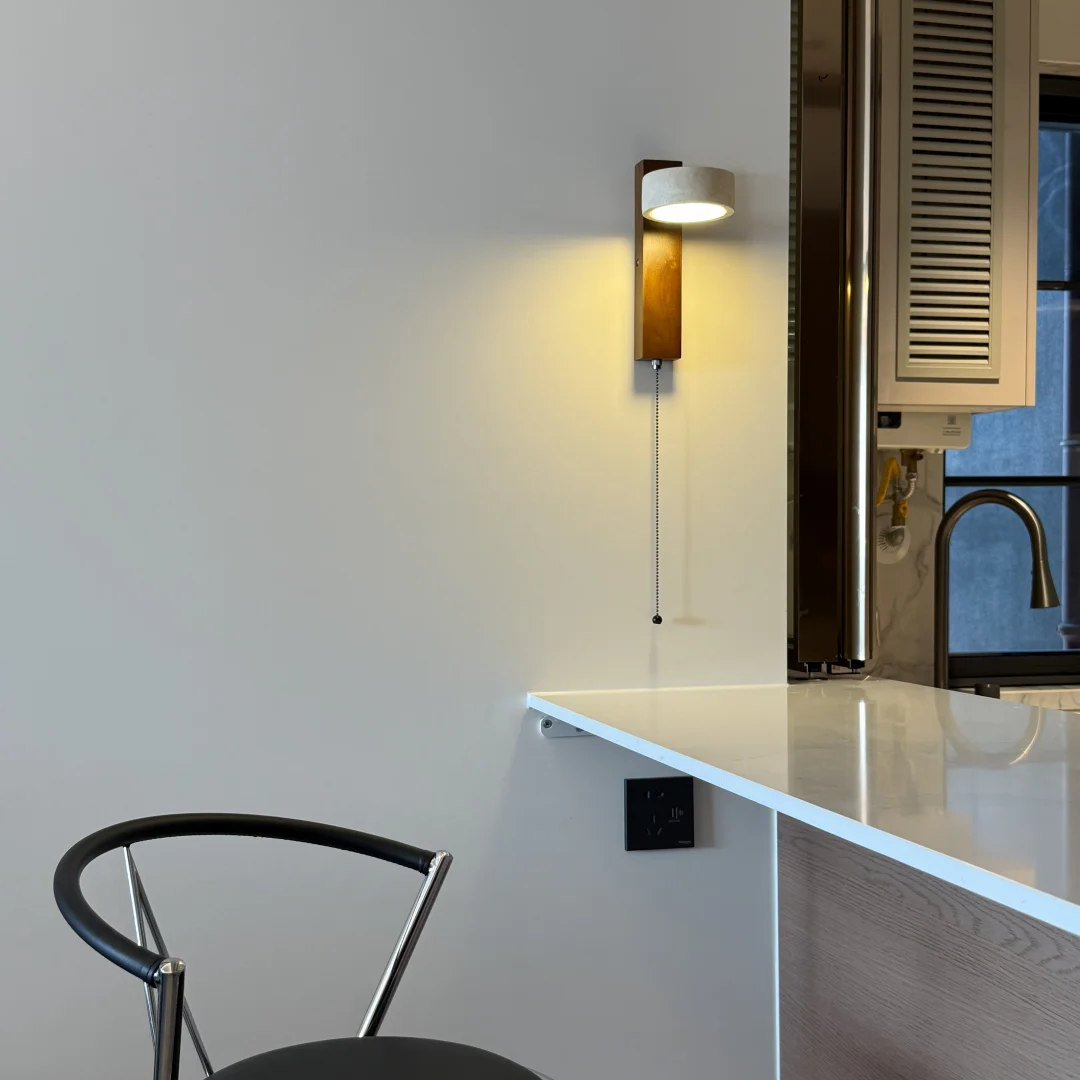A modern wall-mounted sconce with a white cylindrical shade, a vertical wood-look backplate, and a thin cord pull, shown lit on a white wall above a white countertop in a kitchen setting.