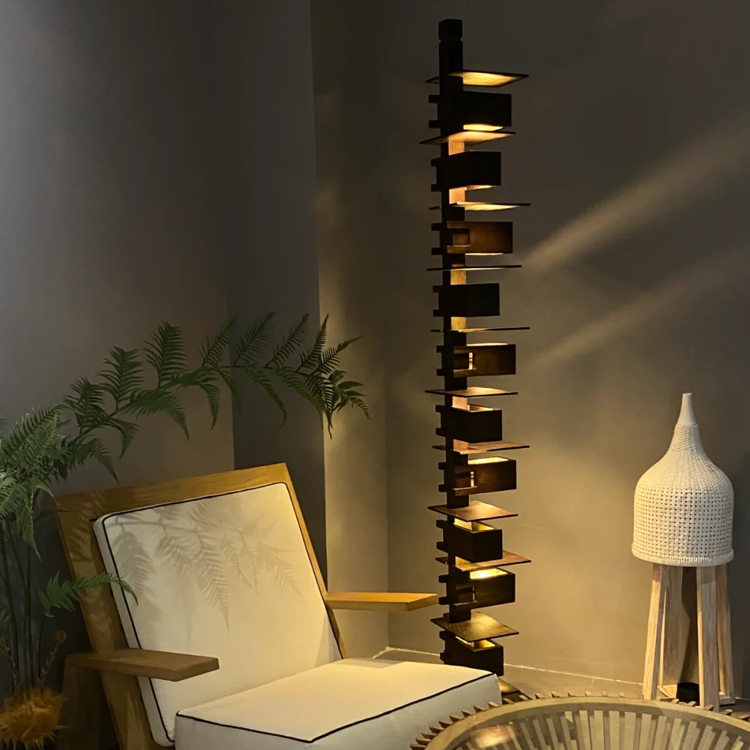 A modern interior scene featuring a tall black floor lamp with multiple illuminated rectangular shelves, a wooden armchair with white cushions, a round slatted coffee table, and a white woven floor lamp against a dark gray wall.