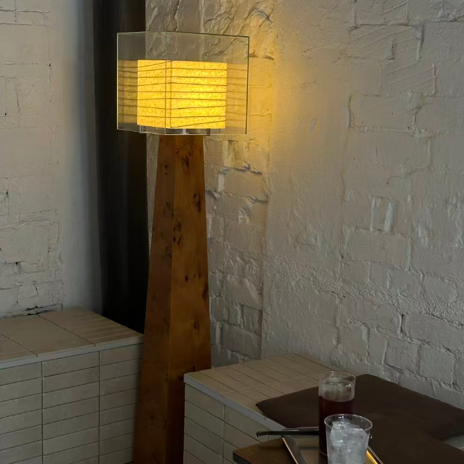 A modern floor lamp with a tall wooden base and a clear rectangular glass shade emitting a warm yellow light, set in a room corner against a white brick wall next to a small table.