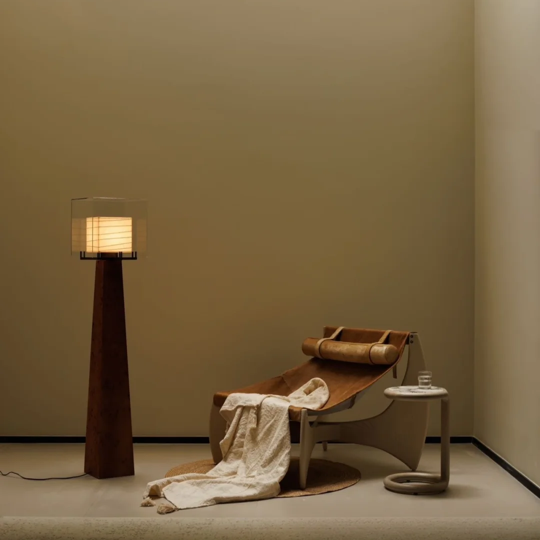 A modern minimalist interior scene featuring a tall dark brown floor lamp with a glowing rectangular shade, a wooden chaise lounge with a cream throw blanket draped over it, and a small round side table against a warm beige wall.