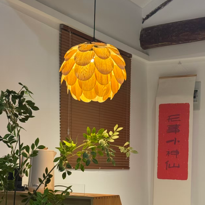 A warm yellow-orange textured pendant light with a layered, scalloped petal-like design hangs in a styled room with a wooden window blind, indoor plants, and a red vertical scroll on the wall.