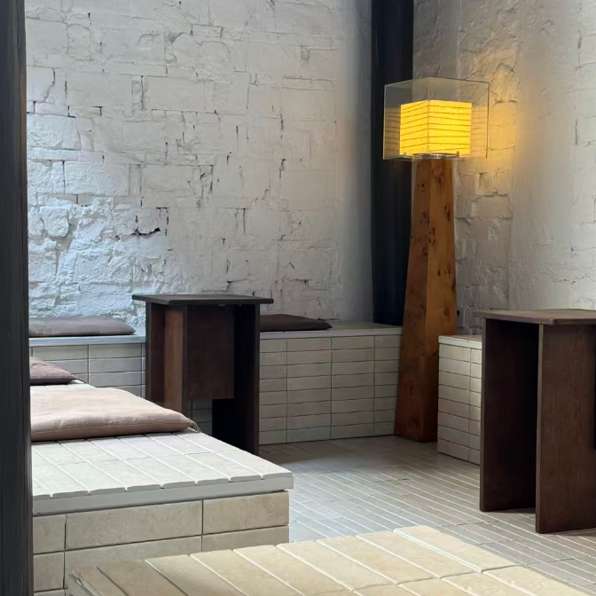 A modern minimalist interior space with white brick walls, low tiled seating platforms, dark wood tables, and a tall floor lamp with a glowing yellow shade.