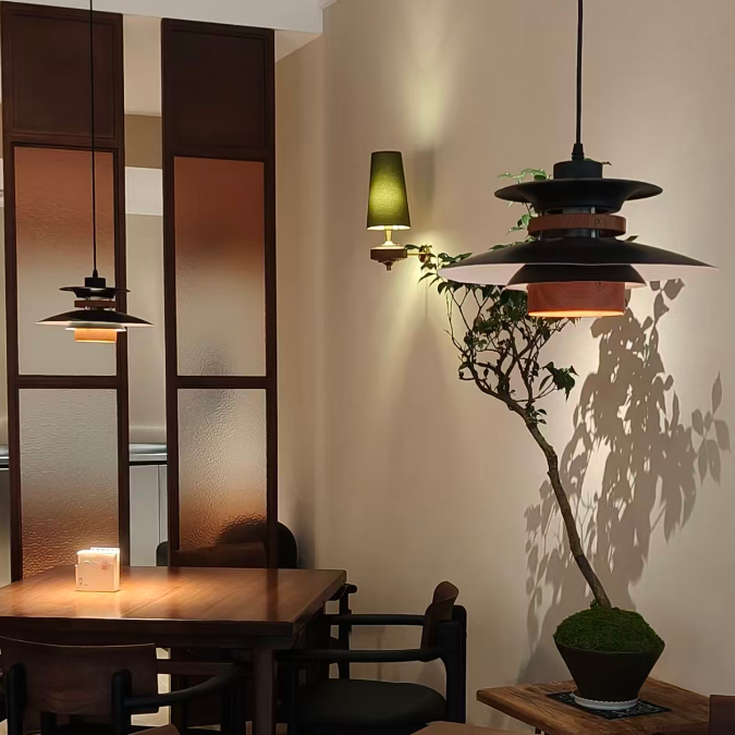 A warm, modern dining room corner featuring a prominent multi-tiered black pendant light with a wooden accent, a matching wall sconce, and a potted bonsai tree casting shadows on the beige wall.