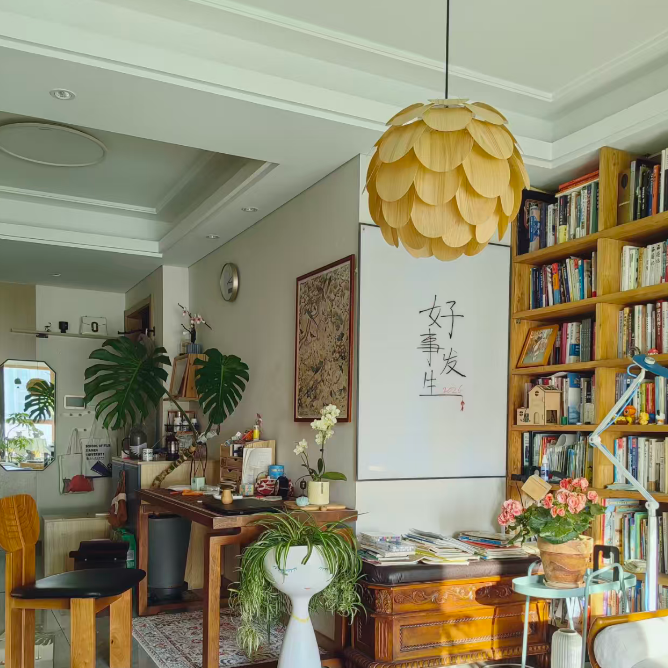 A warm, styled home office or study room featuring a prominent yellow paper-blossom style pendant light hanging from a white ceiling. The room is decorated with a large blue patterned rug, wooden furniture, a bookshelf filled with books, and various potted plants.