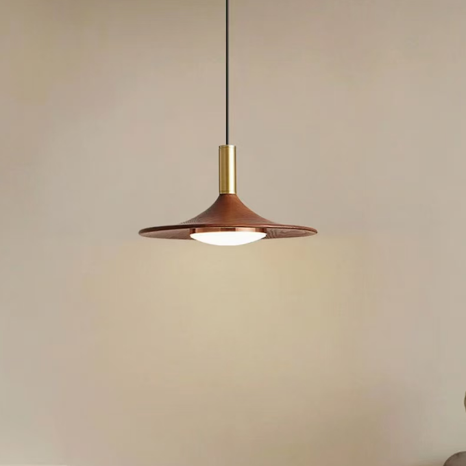 A modern pendant light with a wide, shallow conical shade in a warm brown finish and a brass-toned neck, hanging over a wooden table with stacked plates in a softly lit interior.