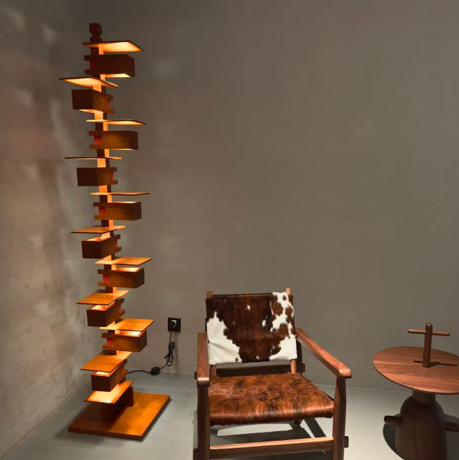 A tall, sculptural wooden floor lamp with multiple tiered shelves emitting warm light stands next to a mid-century modern wooden armchair with a cowhide backrest and seat, in a minimalist room with a concrete wall.