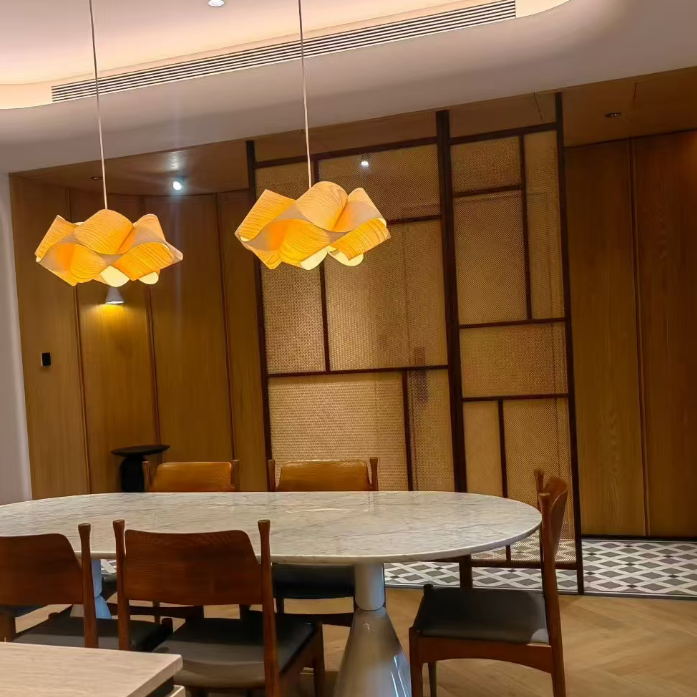 A modern dining room featuring a round white marble dining table surrounded by wooden chairs, with two distinctive wavy yellow pendant lights hanging above. The background consists of a warm wood-paneled wall with geometric patterns.