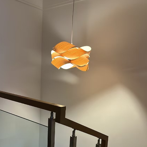 A modern multi-light pendant lamp with warm orange and white wave-like shades hangs over a wooden staircase with a glass railing in a minimalist interior.