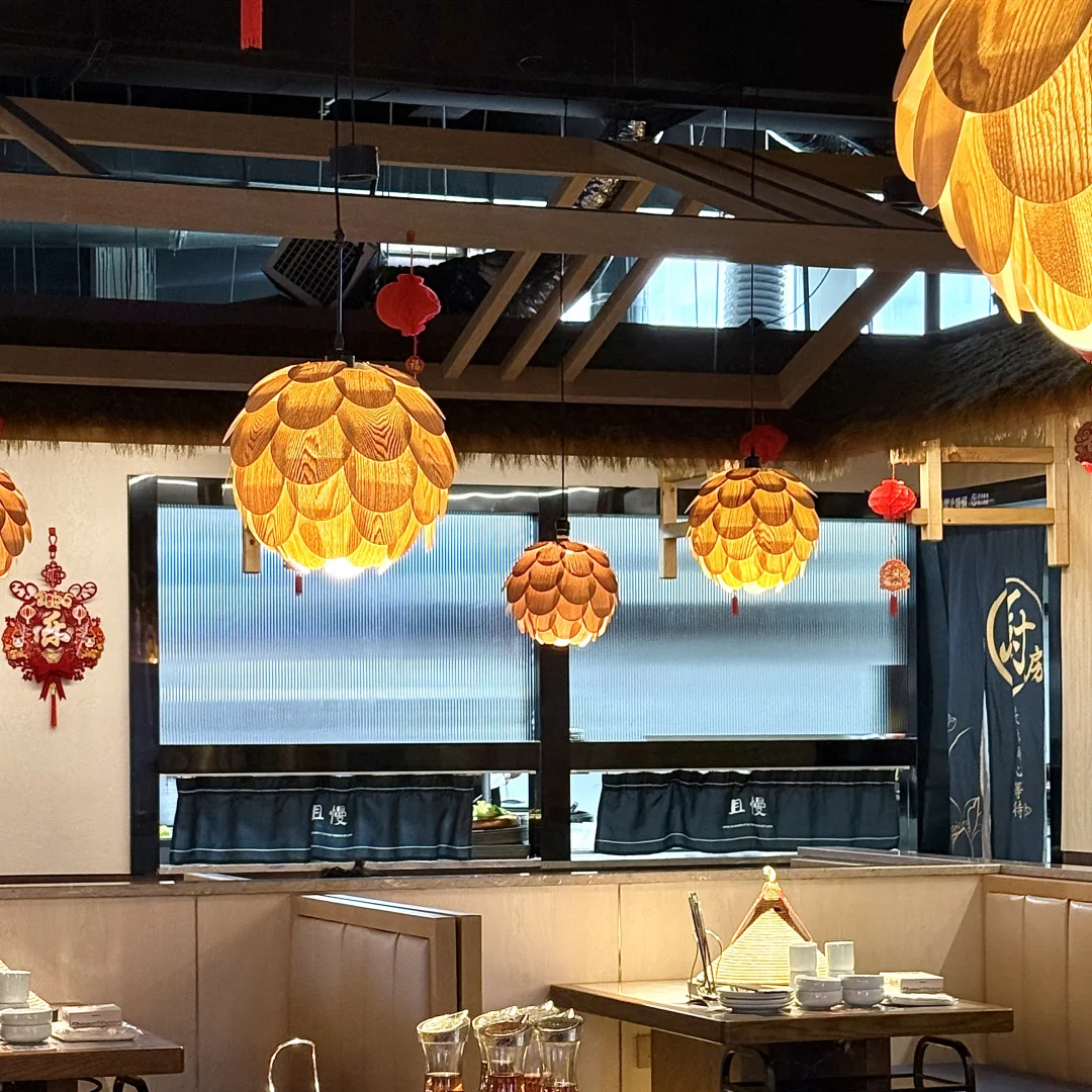 A cozy restaurant interior with round wooden tables, upholstered chairs, hanging textured yellow-orange pendant lights, red lantern decorations, and large windows looking out onto a gray outdoor scene.