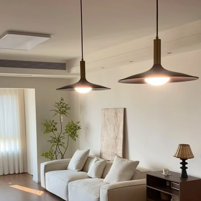 A modern living room featuring two dark bronze conical pendant lights hanging over a light beige sofa. The room has warm neutral tones, a dark wood floor, a side table with a lamp, and a potted plant in the background.
