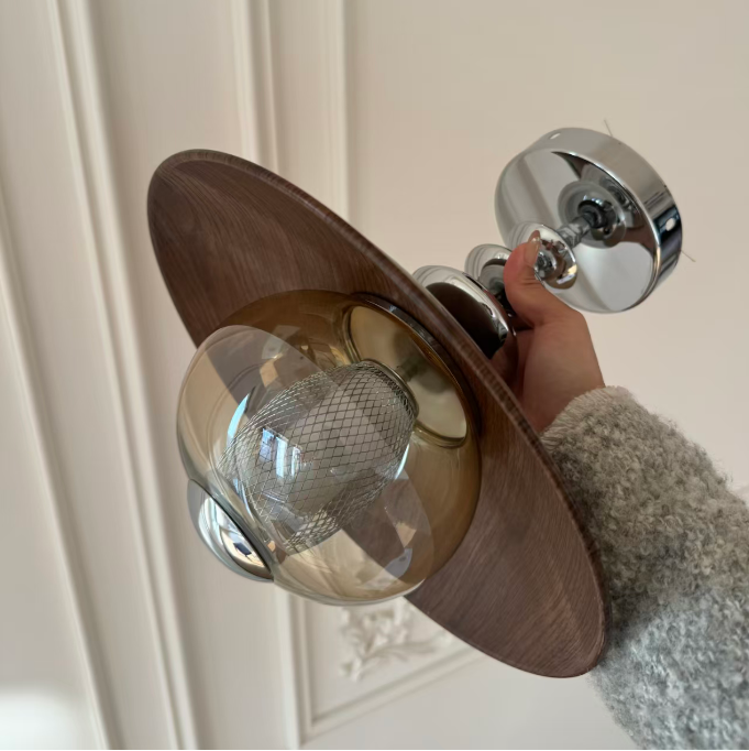 A hand holding a modern wall sconce light fixture with a round dark wood backplate, a translucent amber glass shade, and a polished chrome base against an off-white paneled wall.