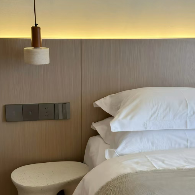A modern bedroom corner featuring a bed with crisp white linens, a textured light beige throw blanket, a sculptural white side table, and a warm wood-paneled headboard with integrated lighting.