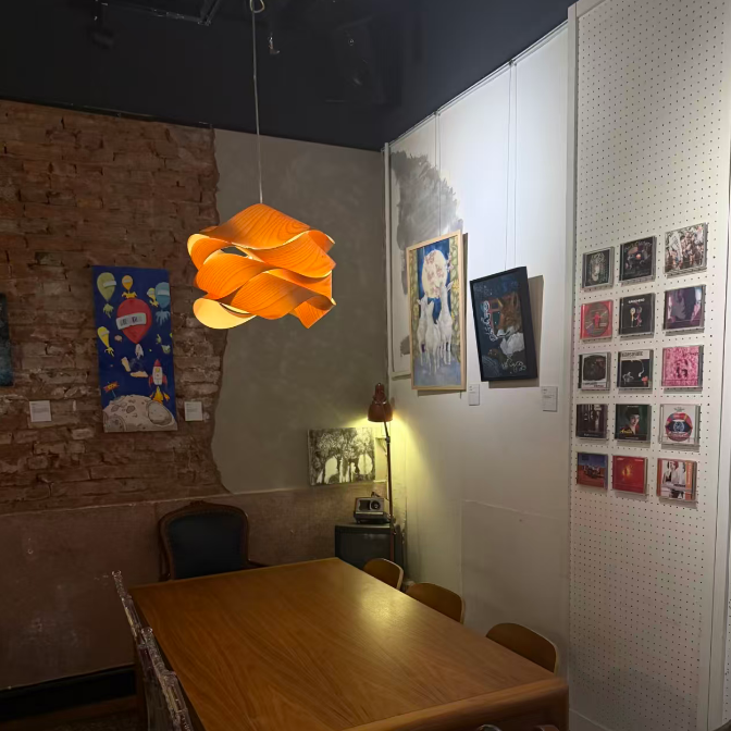 A warm, cozy dining room corner featuring a wooden table and chairs under a distinctive orange, petal-like hanging pendant light. The room has a rustic brick wall on one side and a white pegboard wall with framed art on the other.