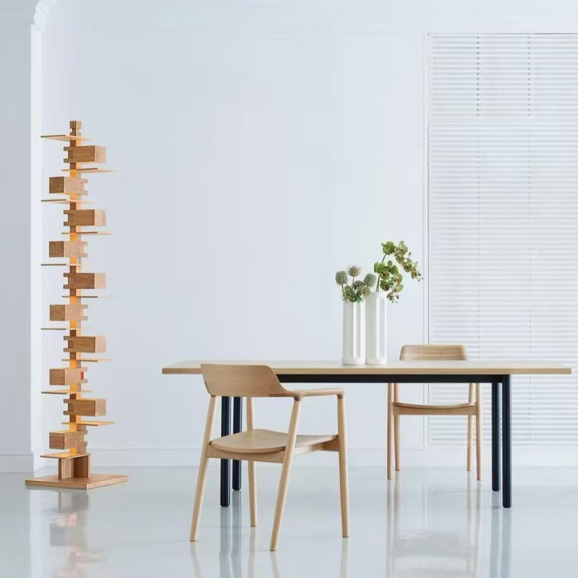 A minimalist dining room scene with a tall wooden geometric shelving unit on the left, a light wood dining table with black legs, and matching wooden chairs. White walls, window blinds, and a glossy white floor complete the setting.