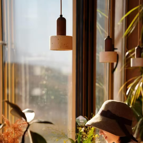 A woman wearing a wide-brimmed beige hat and a cream knit sweater sits at a wooden table by a large window, writing in a sunlit room with hanging pendant lights and potted plants.