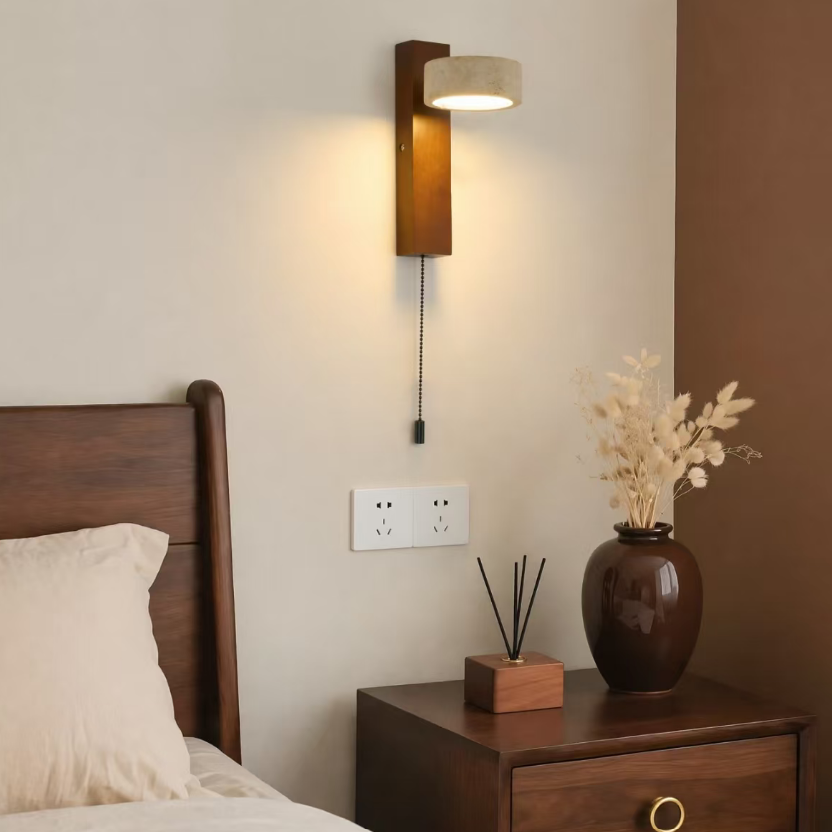 A wall-mounted wooden bedside lamp with a beige drum shade, shown lit in a bedroom setting above a wooden nightstand.