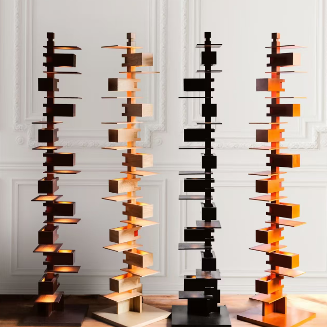 Four modern abstract metal sculpture towers with nested rectangular plates, displayed in copper, bronze, black, and orange finishes against a white paneled wall.