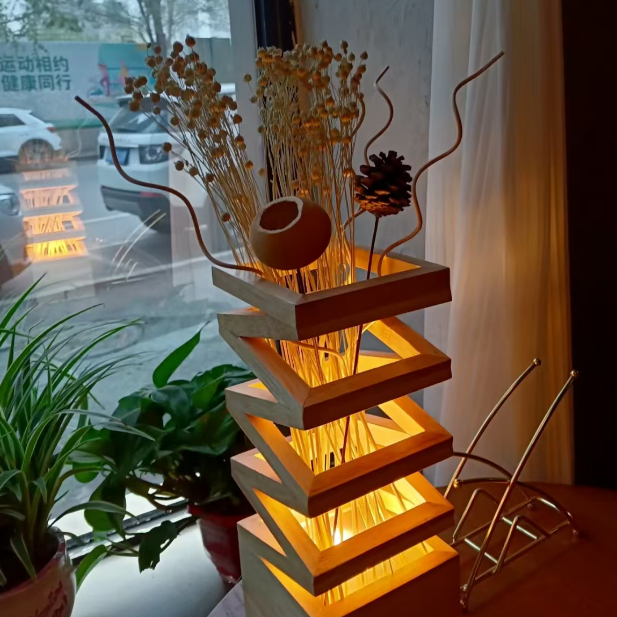 A modern geometric wooden lighted table lamp with dried grass stems and a pinecone, placed on a windowsill next to a potted plant.