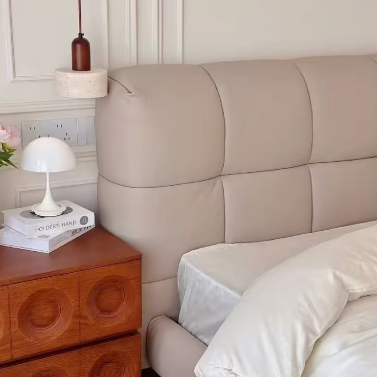 A close-up of a beige upholstered bed headboard and matching bed in a styled bedroom, with white bedding, a wooden nightstand, a white mushroom table lamp, and a hanging light fixture.