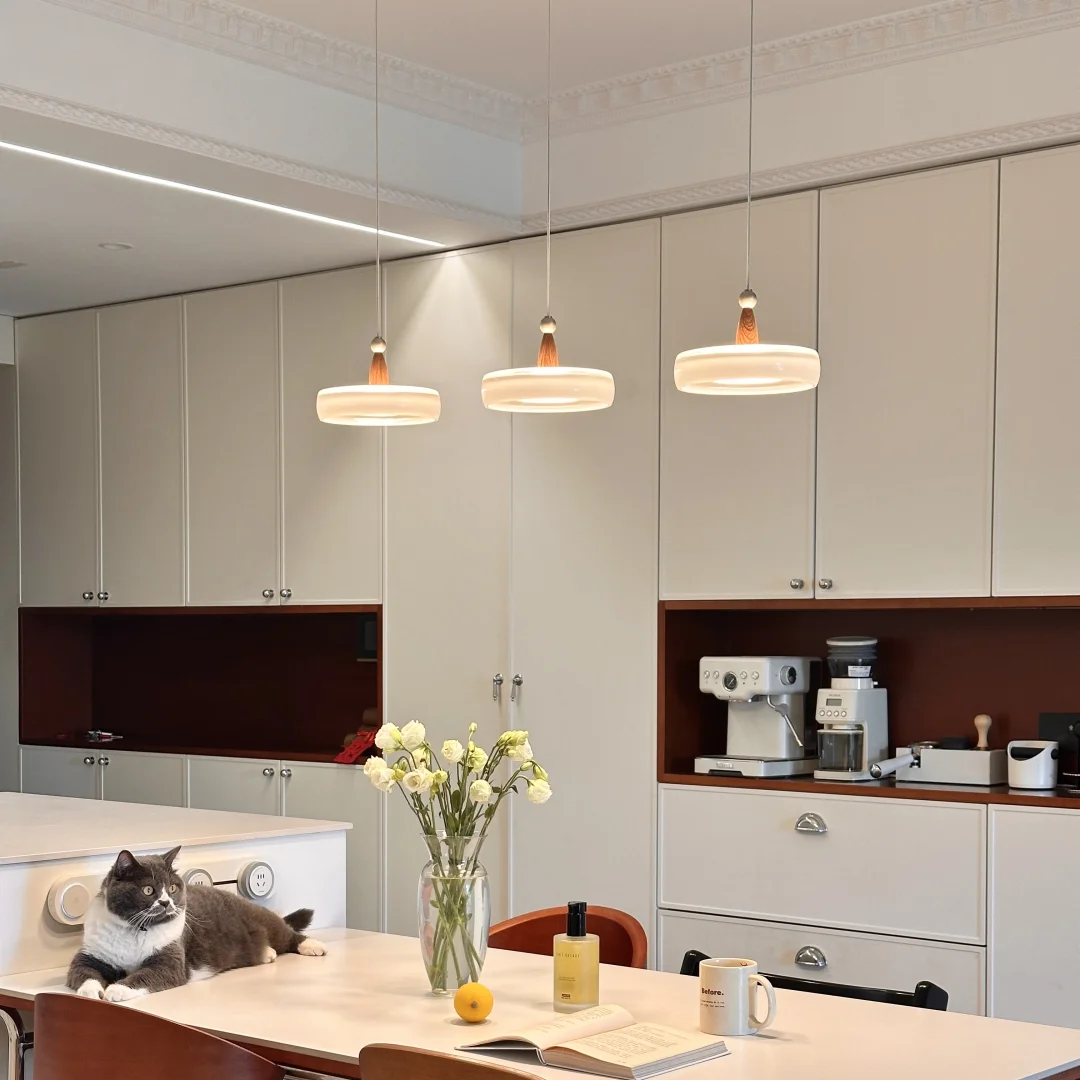 A modern kitchen interior with a long dining table, wooden chairs, pendant lights, and a gray cat lying on the table near a vase of white flowers. Built-in cabinetry, a coffee machine, and open shelving are visible in the background.