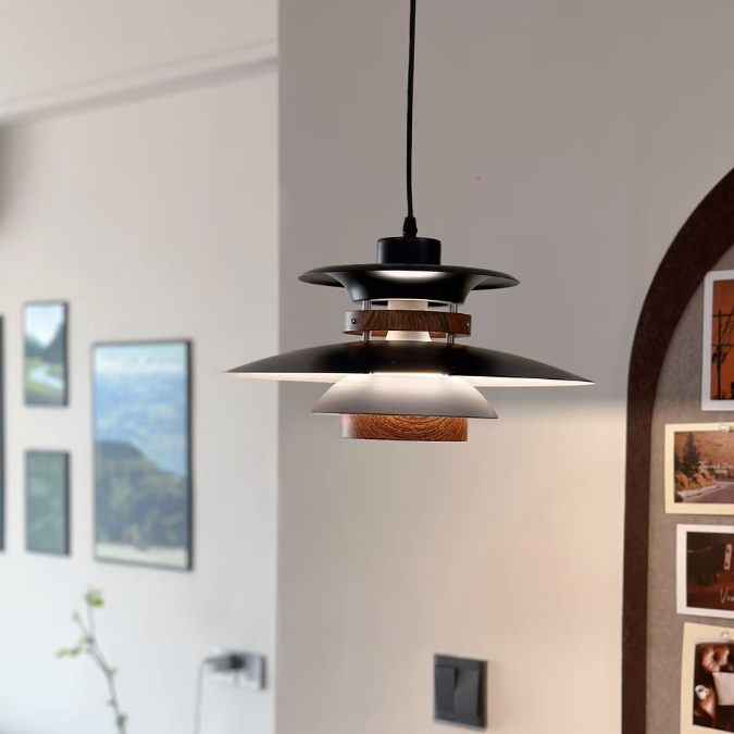 A modern multi-tier pendant light with a black metal finish, warm wood accents, and a white inner shade, hanging in a minimalist, softly blurred interior room.