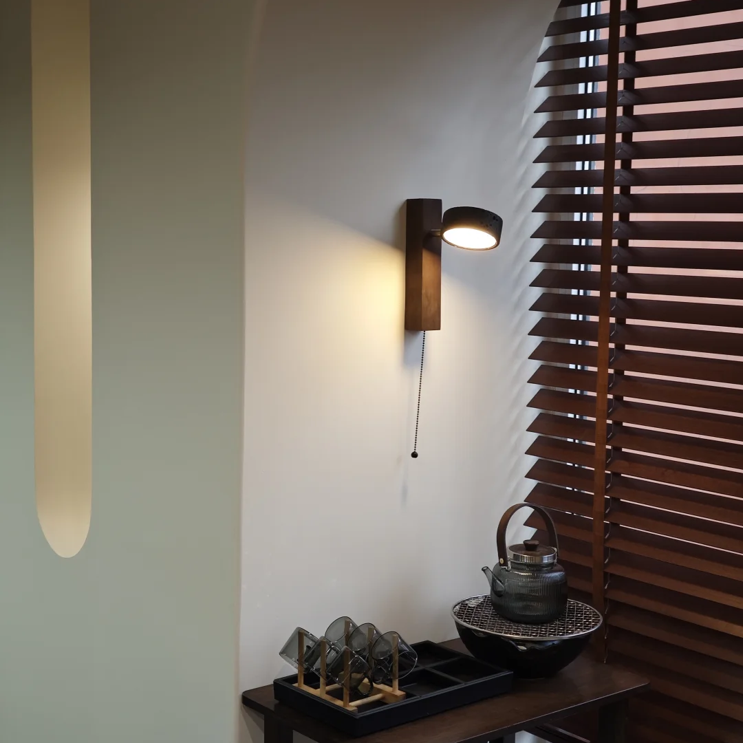 A modern wall-mounted sconce with a dark wood rectangular backplate and a black metal shade, casting a warm light on a cream-colored wall next to dark brown wooden window blinds.