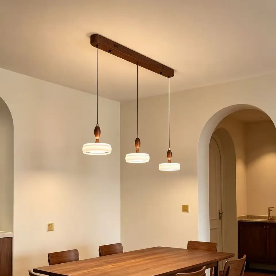 A modern 3-light linear pendant dining chandelier with a dark wood top bar, three hanging glass shades, and warm bulb caps, shown above a wooden dining table in a dining room.