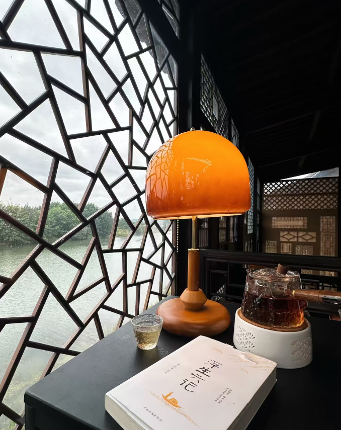 A warm orange dome lamp sits on a dark table beside a glass teapot, a small cup, and a book, set in front of a traditional geometric window overlooking water.