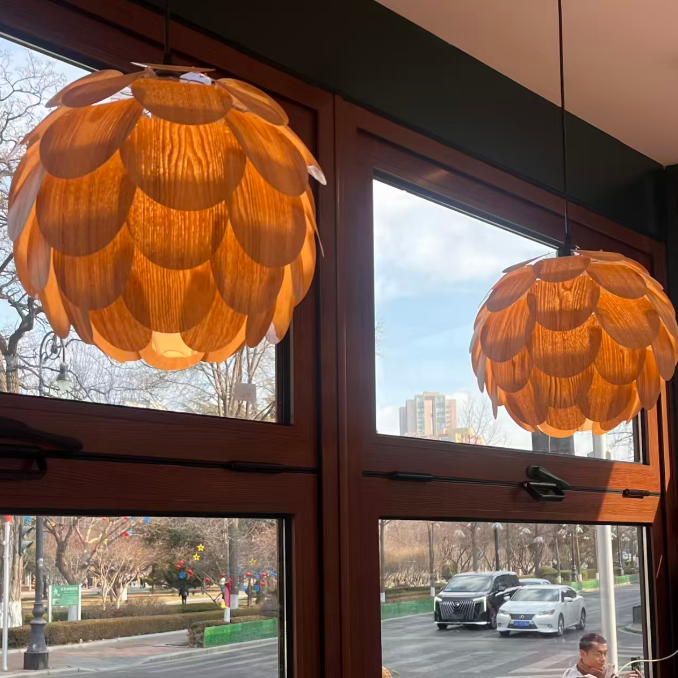 Two large, flower-shaped pendant light fixtures made of overlapping warm orange-brown petals hang on either side of a large window. The window offers a view of a street scene with parked cars and trees outside, and a person is visible in the lower right window area.