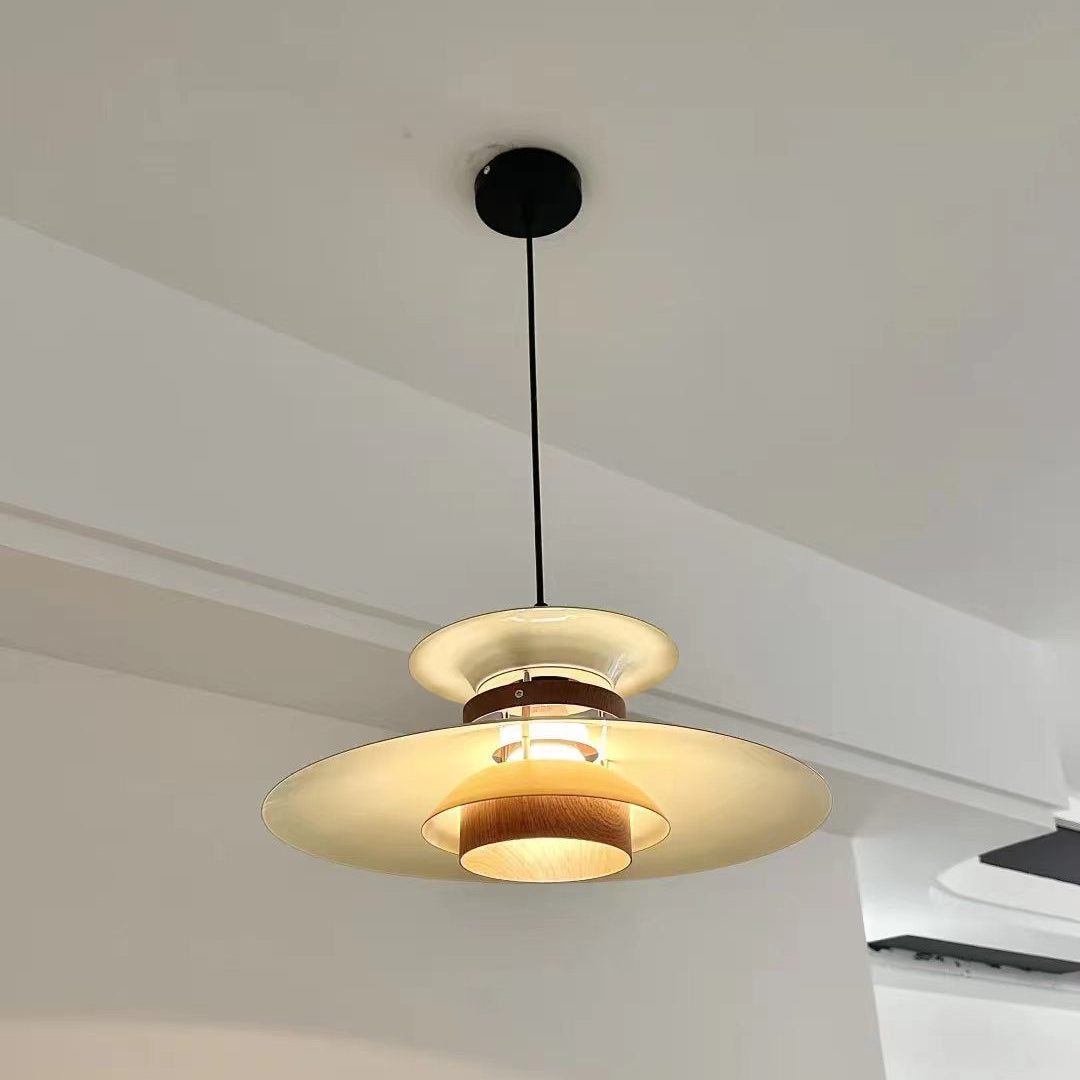 A modern multi-tier pendant light fixture with a black ceiling canopy and cord, featuring cream-colored shades and warm wood accents, shown hanging in a minimalist room with white walls.