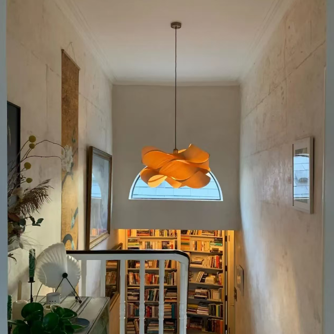 A modern orange flower-shaped pendant light hangs in a narrow hallway above a staircase, with a book-filled library visible below and textured cream walls on both sides.